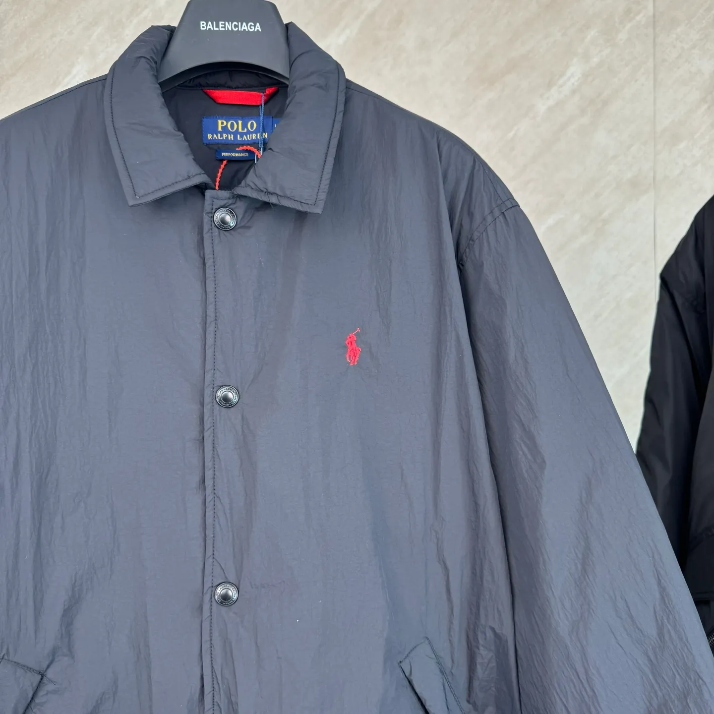 P670 25FW Ralph Lauren red label light cotton jacket (coach jacket) size s m l xl Fabric procurement: a combination of pragmatism and classic texture Material: high-density nylon taffeta Features: density is about 210T-300T, smooth and slightly matte surface, excellent windproof and waterproof performance (with DWR water-repellent treatment), strong wrinkle resistance and easy care, in line with the positioning of American casual "durable and practical".  Color: Classic dark black, using vat dyeing process (dye penetrates into the fiber), the color fastness reaches ISO level 4 or above to avoid fading or color transfer after washing.  2. Lining fabric Material: Lightweight cotton-polyester blended lining (65% Polyester + 35% Cotton) Function: Reduce friction between the fabric and inner clothing, improve wearing smoothness; blended material has both breathability and durability to avoid the stuffy feeling of pure nylon lining.  Version and cutting process Version: Classic Coach Jacket, loose version, slightly dropped shoulder line (shoulder width is 1-2cm wider than the standard version), length to the hips, and slightly tapered straight sleeves, taking into account both freedom of movement and casual style.  Embroidery technology: Polo horse logo on the left chest: flat satin stitch embroidery (satin embroidery), embroidery thread: 100% polyester high color fastness embroidery thread (the color number is consistent with the brand’s standard red color card), stitch density 12-15 stitches/cm, the outline of the horse logo is clear, and the details of the rider and horse are burr-free.  Embroidery positioning: precise positioning through the template (12cm from the neckline and 8cm from the placket) to ensure left and right symmetry and centering.  Topstitching technology: The seams of the body are made of double-needle flat seams (needle pitch 3cm/10 needles), the stitches are smooth and straight, the stitching color is dark black (the same color as the fabric), and the stitch width is 0.5cm, which enhances firmness while forming simple decorative lines.  1. Main body splicing, splicing of the front placket and body: The placket is made of double-layer folding + flat seam splicing, with a thickness of about 0.3cm to ensure that the buttons are not easily deformed when fixed; the edges of the placket are cut with a hot knife to avoid burrs.  Sleeve panels are spliced ​​to the body: the armholes are spliced ​​with round-head keyhole machine seams + flat seams. After splicing, the armholes have a natural curvature and no sense of tightness during movement; the height of the sleeves is about 8-10cm, which is in line with the loose characteristics of casual jackets.  Main button: black urea resin button, matte surface treatment, buttonholes are hand-made keyholes (6 strands of cotton thread, keyhole density 8 stitches/mm), the edges of the eyelets are smooth and without burrs.  Collar buttons: The first button on the collar is a hidden button (diameter 12mm), which prevents the collar from opening and improves the neatness of wearing.